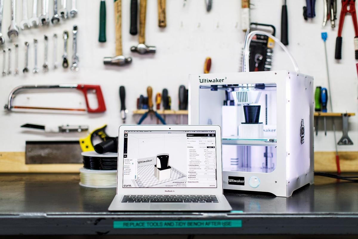 Werkbank met gereedschap aan de wand, een laptop met een 3D-model en een printer die het onderdeel print